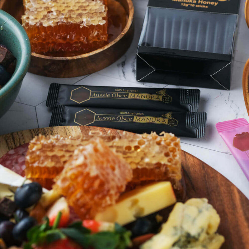Aussie Qbee Manuka honey sachets displayed with honeycomb and assorted fruits on a wooden platter.