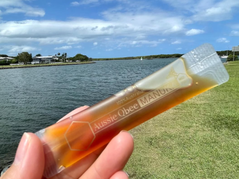 Hand holding a stick of Aussie Bee Manuka honey against a scenic background with water and sky.